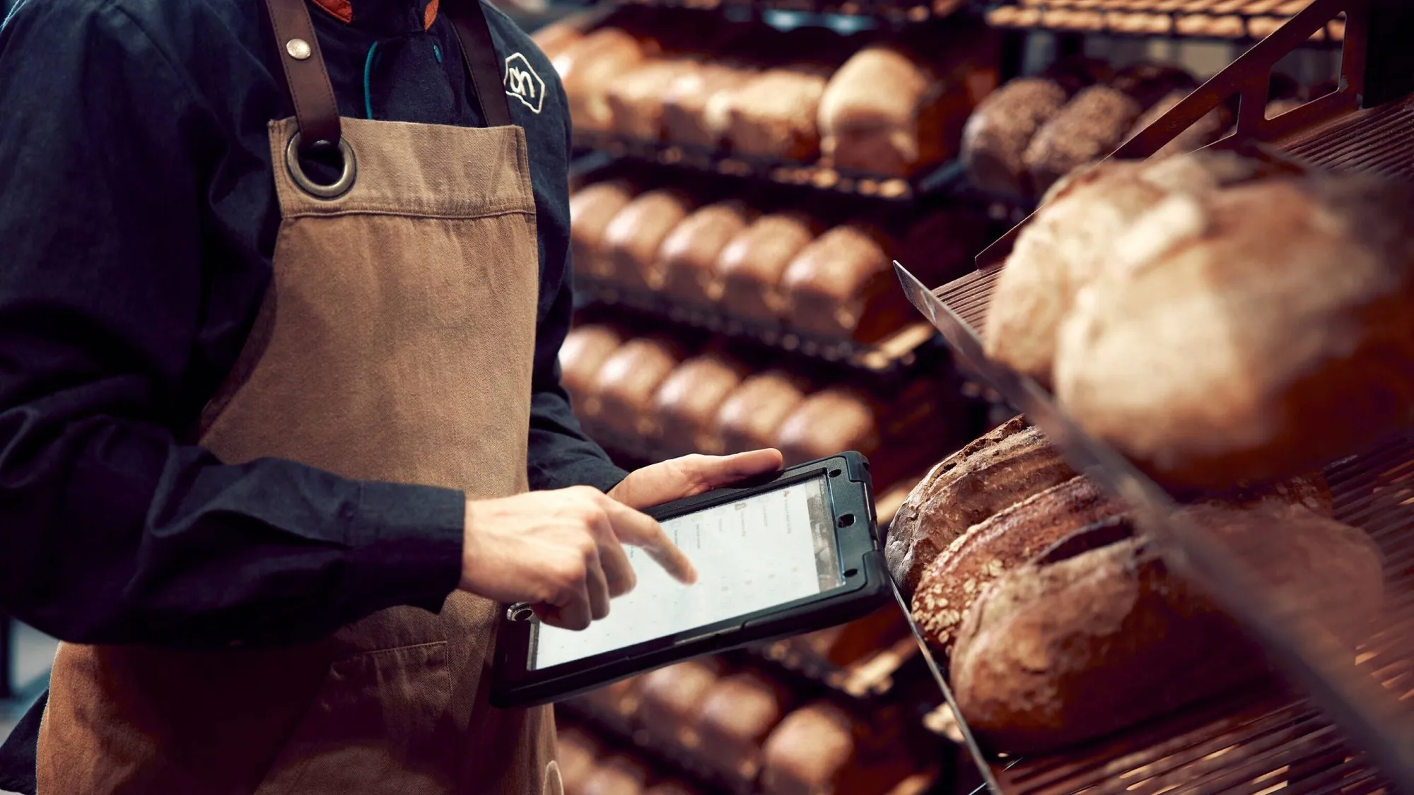 A bakery employee in an Albert Heijn apron tapping an iPad in a rugged case, next to shelves full of bread