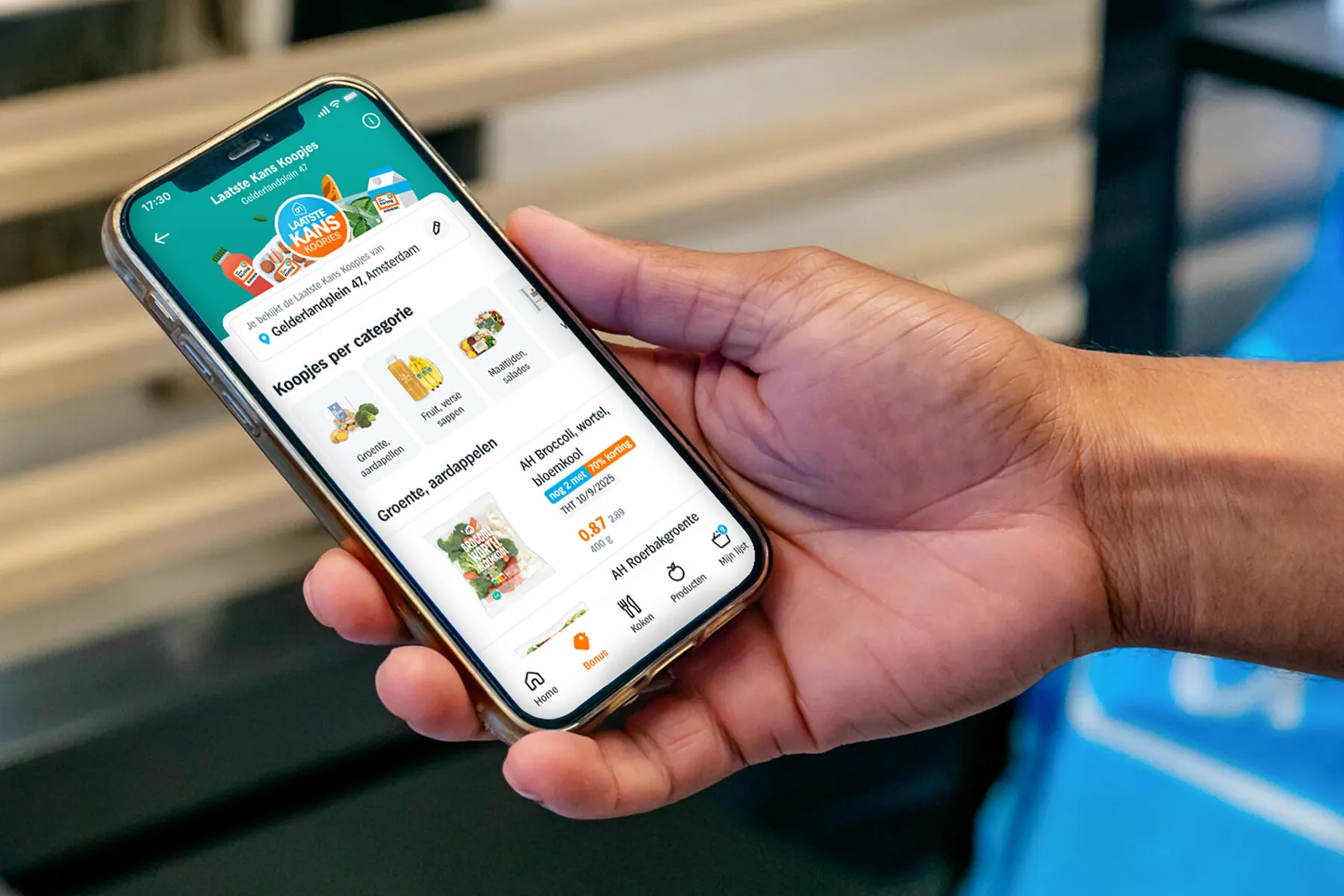 A hand holding a phone with the Laatste Kans Koopjes screen of the Albert Heijn app, showing discounted products per category for a store, such as a bag of vegetables with 70% off