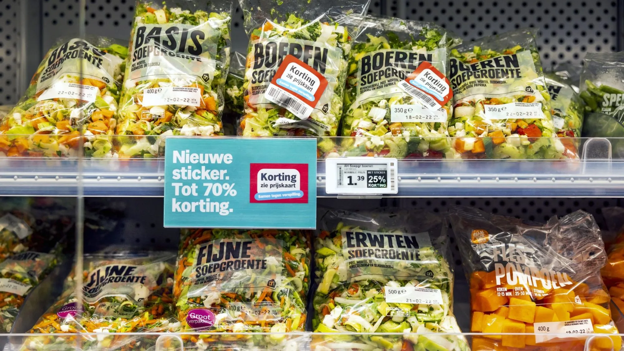 Bags of soup vegetables on a supermarket shelf, two with a red 'Korting, zie prijskaart' sticker, next to a sign announcing the new sticker with up to 70% off and an electronic shelf label showing 25% off